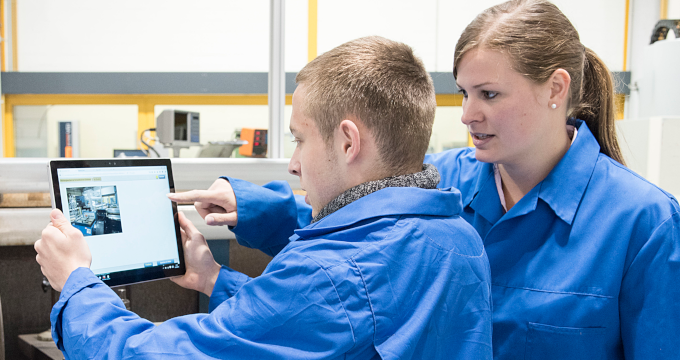 Auszubildender und Auszubildende in blauer Arbeitskleidung in fabrikähnlicher Umgebung. Der Auszubildende hält ein Tablet hoch, auf das beide blicken.